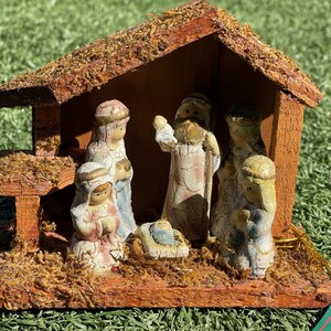 May include: A wooden nativity scene with figures inside a rustic shelter. The figures are painted in muted tones, depicting the traditional Christmas story. The roof is covered in moss, and the scene is set on a bed of dried grass.