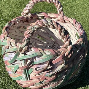 May include: A round woven basket with a braided handle. The basket is made of interwoven strips of natural and dyed materials in shades of green, pink, and brown. The basket is sitting on green grass.