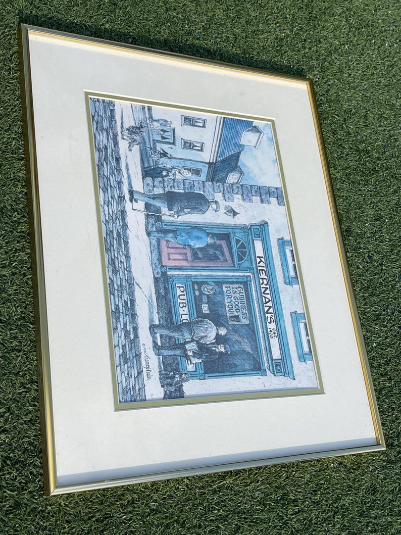 May include: Framed print of a pub scene with figures outside. The pub's sign reads "Kiernan's Pub" and "Guinness is good for you." The artwork is in a gold frame with a white mat, set against a green background.