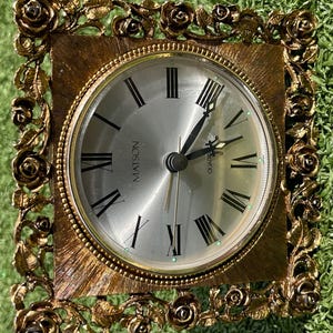 May include: A square, gold-toned clock with a silver face and black Roman numerals. The clock face reads "MATSON" and "QUARTZ". The frame is decorated with roses. The clock is likely vintage or antique.