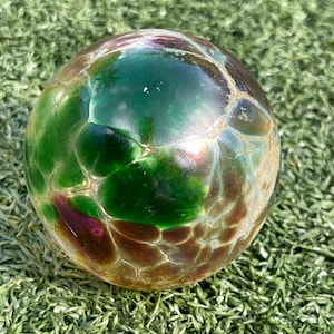 May include: A decorative glass sphere with a mosaic-like pattern. The sphere features shades of green, brown, and hints of purple. The glass has a textured appearance, and the sphere is resting on a bed of green artificial grass.