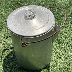 May include: A vintage metal coffee pot with a glass knob on the lid and a wire handle. The pot is silver-colored and appears to be made of aluminum. The pot is sitting on a green surface.