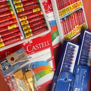 Op de afbeelding: Een verzameling kunstbenodigdheden waaronder twee witte plastic trays gevuld met Faber-Castell oliepastels, een rode Faber-Castell doos, een verpakking met zes ArtSkills penselen en twee blauwe dozen met Bazic oliepastels met twaalf kleuren.