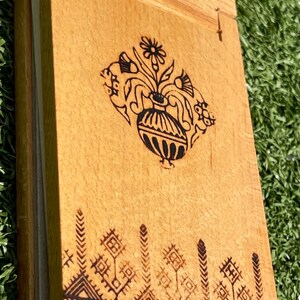 May include: Wooden notebook with a decorative floral and vase design, and geometric patterns. The notebook is made of light-colored wood and has a hinged cover. The design is burned into the wood, creating a dark brown contrast.