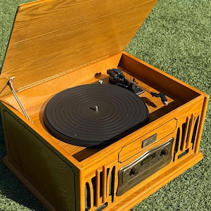 Puede incluir: Un tocadiscos de madera con tapa abatible, con un plato giratorio negro y un panel de control plateado. La unidad tiene un acabado marrón dorado cálido y está sobre un fondo de hierba verde. Un dispositivo de audio de estilo vintage.