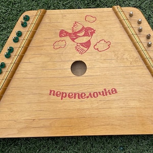 May include: A wooden zither featuring a light brown finish, metal strings, and green tuning pins. The instrument displays a red graphic of a bird with clouds and the Cyrillic text "перепелочка" printed in red ink on the front soundboard.