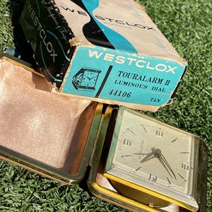 May include: Vintage Westclox Touraalarm II luminous dial travel alarm clock in its original box. The clock has a tan face and a gold-tone case. The box is blue, black, and white with the Westclox logo and product information.