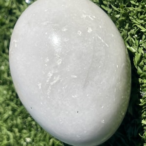 May include: A smooth, oval-shaped, white stone. The stone has a slightly glossy surface and subtle variations in color. The stone is resting on a bed of green artificial grass. The stone appears to be a decorative item or a natural element.