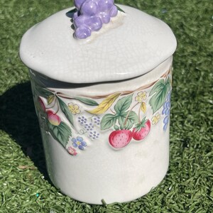 May include: A ceramic jar with a lid, decorated with a fruit and floral design. The lid is topped with a cluster of purple grapes. The jar features strawberries, cherries, and blueberries.