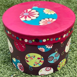 May include: A round, decorative gift box with a magenta lid and a dark brown base. The lid features a teal ornament design with white and pink floral accents. The base is adorned with colorful ornament illustrations, perfect for holiday gifting.