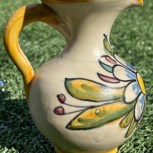 May include: A small, cream-colored ceramic pitcher with a yellow handle, rim, and base. The pitcher is decorated with a floral design featuring blue, green, and pink accents. The pitcher is likely used for serving beverages.