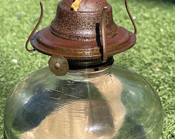 May include: A vintage oil lamp with a clear glass globe and a rusty brown metal top. The lamp has a round, bulbous shape and is designed to sit on a flat surface. The top features a wick and a small handle.