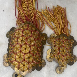 May include: Two decorative ornaments made of gold-colored coins, intricately woven with red and gold threads. Each ornament has a tassel of gold and red cords. The coins are arranged in a geometric pattern, creating a unique and eye-catching design.