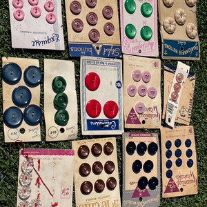 May include: A collection of vintage buttons in various colors, including pink, purple, green, red, brown, and blue. The buttons are arranged on their original cards, with some cards featuring text like "Exquisit" and "Le Chic".