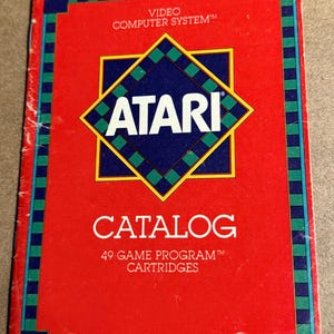 May include: A vintage Atari catalog with a red background and a blue and green border. The Atari logo is prominently displayed in the center, with the words "CATALOG" and "49 GAME PROGRAM CARTRIDGES" below.