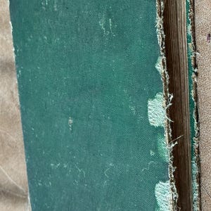 May include: An antique book with a distressed green cover. The edges of the cover are frayed, revealing the pages within. The book shows signs of age and wear, with a textured appearance.