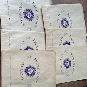 May include: Several beige paper bags are stacked, each printed with a purple sun design and the words "PROVENANCE GALLERIES." The text "Philipsburg, St. Maarten, Marigot St. Martin, Naples, Florida" is also printed on the bags.