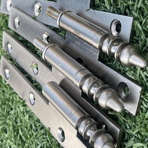 May include: Close-up of several silver-toned metal hinges with decorative, rounded finials. The hinges are laid out on a bed of green grass. Each hinge has a rectangular plate with multiple screw holes.