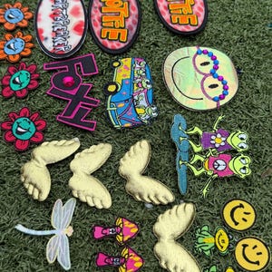 May include: A collection of colorful embroidered patches on a green surface. Designs include flowers, smiley faces, wings, mushrooms, a dragonfly, and text that reads "Hottie" and "Heartbreaker".