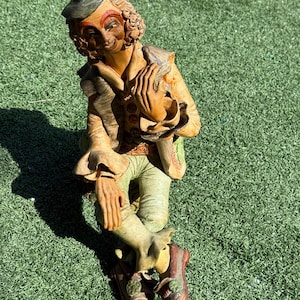 May include: A whimsical figurine of a seated jester with a black hat, curly hair, and a smiling face. The jester wears a tan jacket, green pants, and brown shoes. The figurine is set against a green background.
