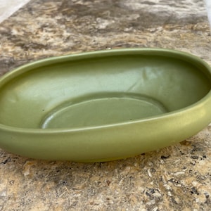May include: A light green, oval-shaped ceramic planter. The planter has a smooth, rounded rim and a slightly recessed interior. The color is a muted, earthy green, and the overall design is simple and modern. The planter is suitable for small plants or succulents.