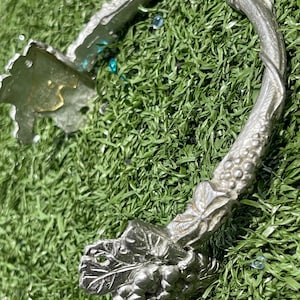May include: Silver-toned wine bottle opener with a decorative grape and leaf design. The opener has a curved shape and is made of metal. The design includes detailed grape clusters and leaf accents, adding a touch of elegance.