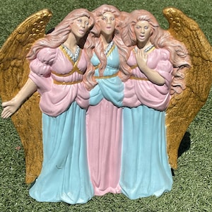 Peut inclure: Une figurine décorative représentant trois personnages debout avec de longs cheveux et de grandes ailes dorées. Les personnages portent des robes fluides rose pastel et bleu clair, avec des ceintures dorées et des détails argentés au cou.