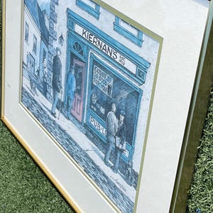 May include: Framed print of an Irish pub, "Kiernan's", with the text "Guinness is good for you". The artwork depicts a street scene with figures outside the pub, rendered in blue, white, and gray tones. The frame is gold.