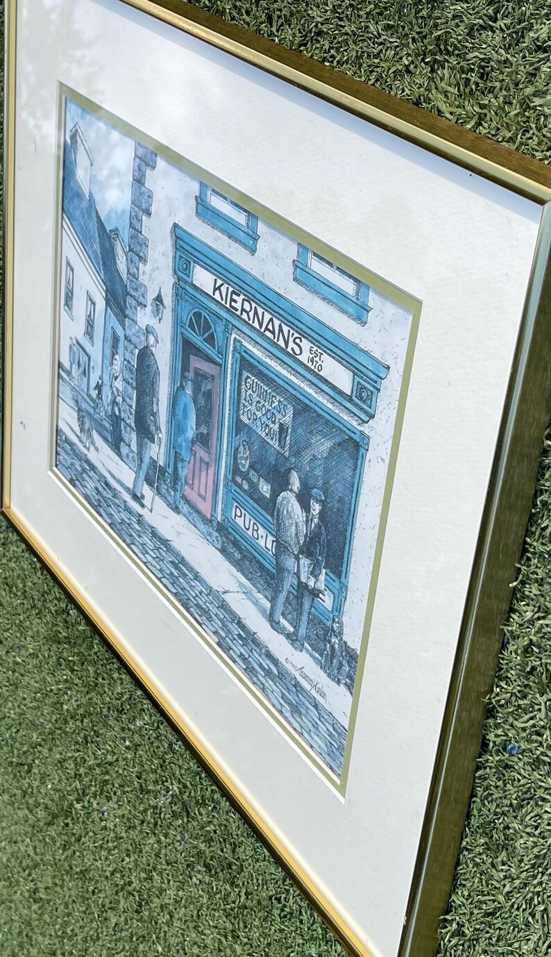 May include: Framed print of an Irish pub, "Kiernan's", with the text "Guinness is good for you". The artwork depicts a street scene with figures outside the pub, rendered in blue, white, and gray tones. The frame is gold.