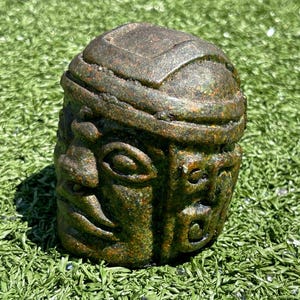 May include: A weathered, stone-like sculpture of a stylized human head with two faces. The sculpture is primarily dark green and brown, with detailed facial features and a band around the head. The sculpture is set against a backdrop of green artificial turf.
