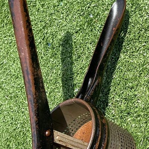 May include: An antique potato ricer with a perforated bowl and two long handles. The metal tool has a dark brown and black finish, showing signs of age and use. The ricer is designed for pressing cooked potatoes.