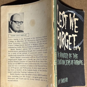 May include: Open book with a black and white photo of Rabbi Leivy Smolar and text. The book's title is "Lest We Forget... The Murder of the Six Million Jews of Europe" by Leivy Smolar, part of the Judaism Pamphlet Series.