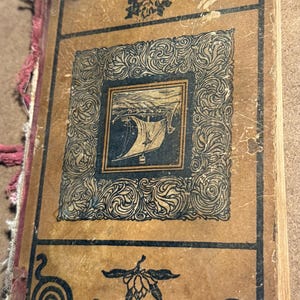 May include: Vintage journal with a worn, tan cover featuring a central framed illustration of a sailboat. The frame and corner designs are dark blue, with decorative swirls. The journal's spine has a pink ribbon.