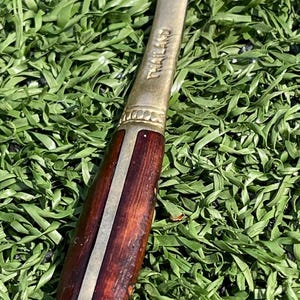 May include: A vintage spoon with a brass bowl and handle. The handle is made of dark wood with a brass accent and the word "THAILAND" is engraved on the handle. The spoon is set against a backdrop of green.