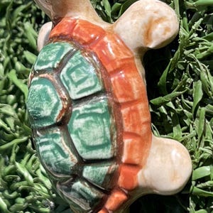 May include: A ceramic turtle figurine with a green and orange shell. The turtle has a tan head, legs, and tail. The shell is detailed with a geometric pattern. The figurine is placed on a bed of green artificial grass.