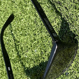 May include: Black sunglasses with the brand name "Vuarnet" printed on the arm. The sunglasses are resting on a bed of green grass. The model number "900" is printed on the left arm.