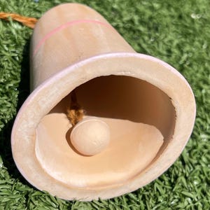 May include: A light brown bell with a pink accent line and a light pink rim. A small, round, light brown clapper hangs inside the bell. The bell is suspended by a brown rope.