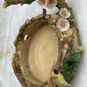 May include: A decorative wall-mounted bird feeder with a rustic, tree-like design. It features a central bowl, adorned with pink flowers, green leaves, and a hummingbird. The feeder is made of a light brown material.