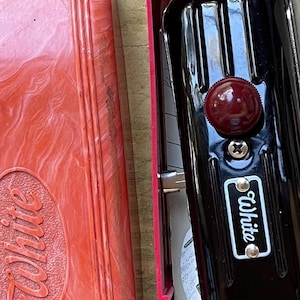 May include: A vintage black sewing machine with a red knob and the name "White" on a white label. The machine is in a red case with the name "White" embossed on the lid. The case has a marbled design.