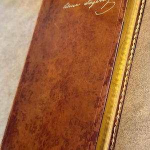 May include: A vintage, leather-bound book with a rich, brown cover. The book's pages are gilded, and the spine is adorned with gold detailing. The cover features elegant, gold-colored script. The book's aged appearance suggests it is an antique.