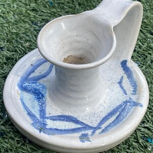 May include: A handmade ceramic candle holder with a white base and blue abstract leaf design. The candle holder has a circular base and a curved handle. The interior of the candle holder is a light brown color.