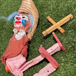 May include: A vintage clown puppet with a straw hat, blue hair, and a painted face with red lips and blue teardrops. The clown wears a red shirt and pink pants. The wooden control cross and strings are visible, suggesting a marionette.