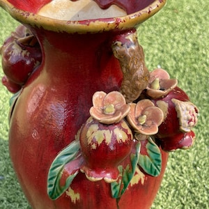 May include: A red ceramic vase with a wide, ruffled rim and a textured surface. The vase is adorned with sculpted pomegranates, flowers, and green leaves. The rim has a yellow-brown trim. The vase is likely for decorative purposes.
