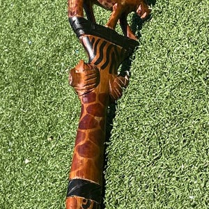 May include: A wooden walking stick with carved animal figures. The handle has a curved design with black and brown patterns. The shaft features a giraffe pattern and a carved lion. The stick is made of brown wood.
