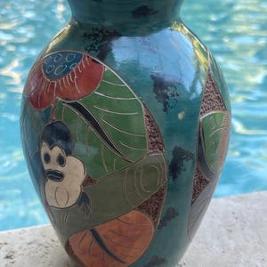 May include: A teal ceramic vase with a detailed design. The vase features a monkey, leaves, and floral patterns in red, green, and brown. The vase has a glossy finish and a wide opening.