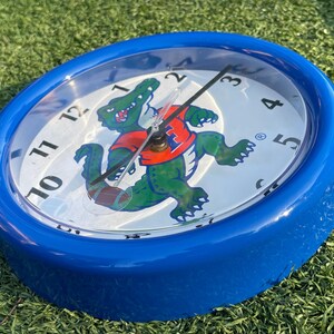 May include: Blue oval clock with a white face featuring black numerals and hands. A green alligator mascot wearing an orange shirt with a blue "H" logo is in the center. The clock is a sports-themed item.