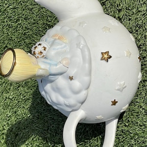 May include: White ceramic teapot with a whimsical design. The teapot features a sleeping angel figure and gold star accents. The handle and spout are curved, and the teapot is set against a green background.