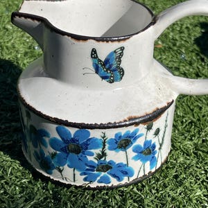 May include: White ceramic pitcher with a butterfly and blue floral design. The pitcher has a spout and a handle. The rim and edges are outlined in brown. The blue flowers are painted around the base of the pitcher.