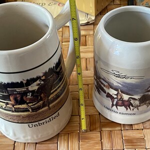 May include: Two white ceramic beer steins with handles. One features a rodeo scene with the word "Unbridled". The other stein has a winter scene with the word "Stetson" and "Limited Edition". The steins are approximately 5 inches tall.