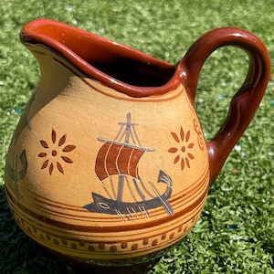 May include: A small, tan ceramic pitcher with a brown handle and rim. The pitcher is decorated with a ship and floral designs in brown and red. The pitcher is sitting on a green surface.